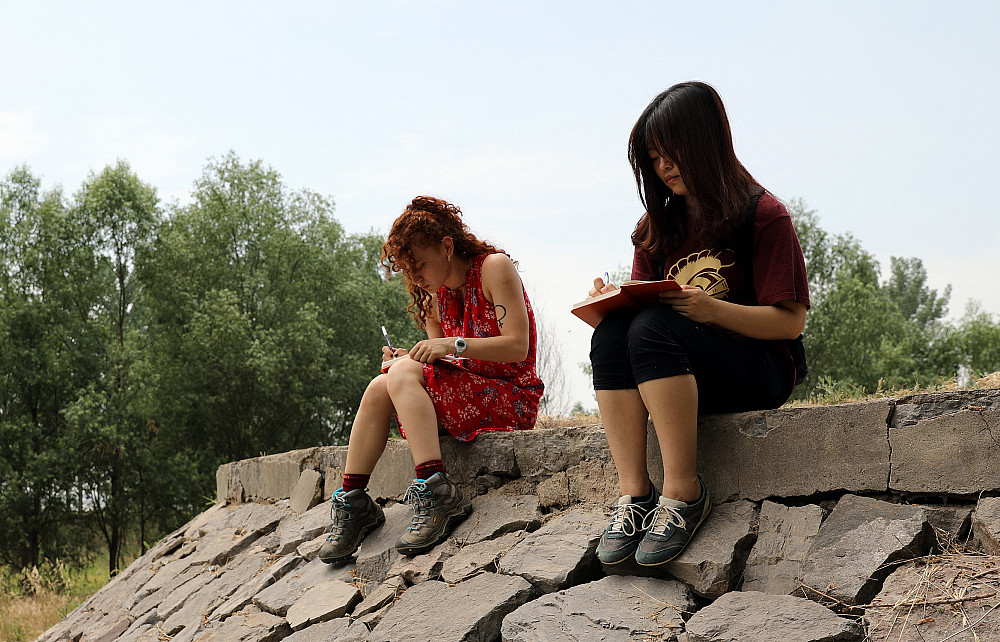 Two students sit on a mound of rocks, writing.