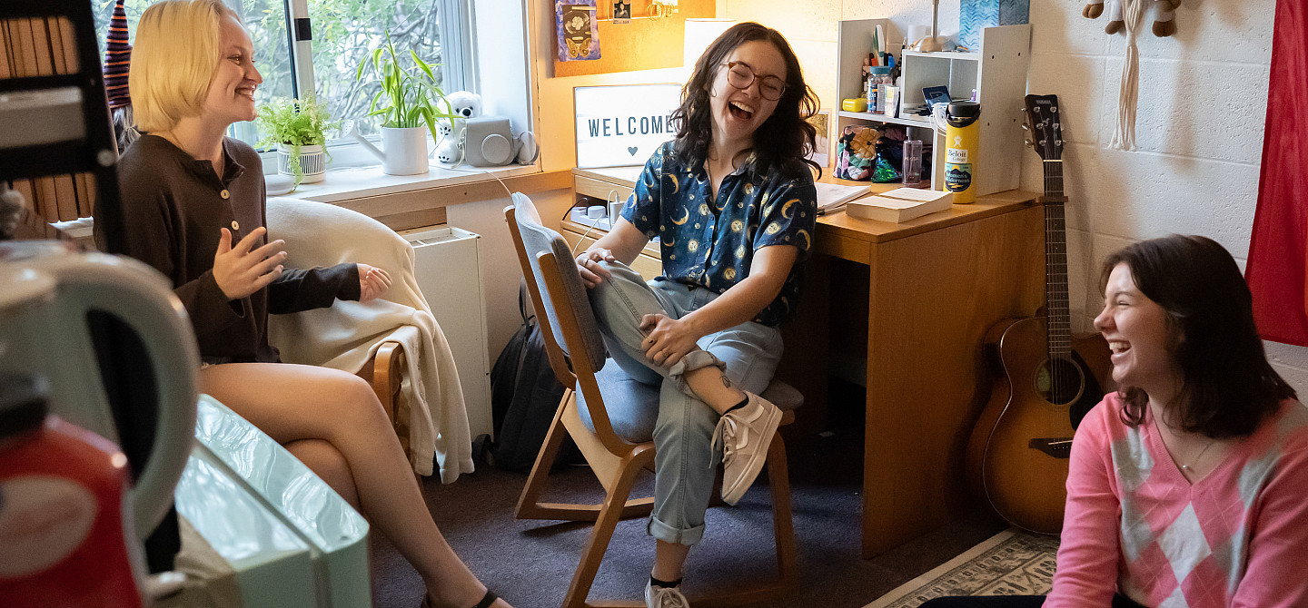 Three students hanging out together in a dorm room, laughing together.