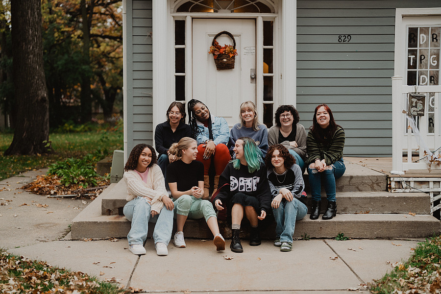 Members of the Theta Pi Gamma sorority sit together on the porch of their house, smiling and laughing.