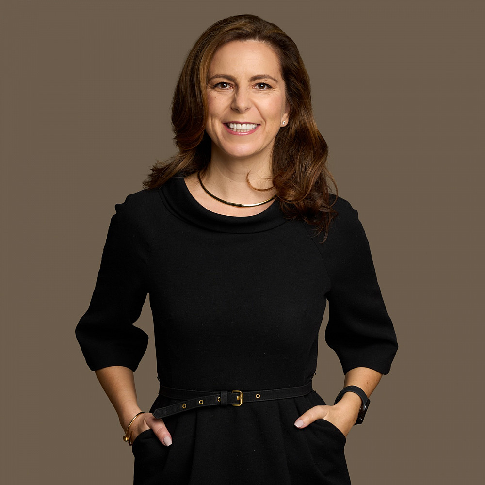 A woman in a dark colored dress with her hands in her pockets smiles at the camera.