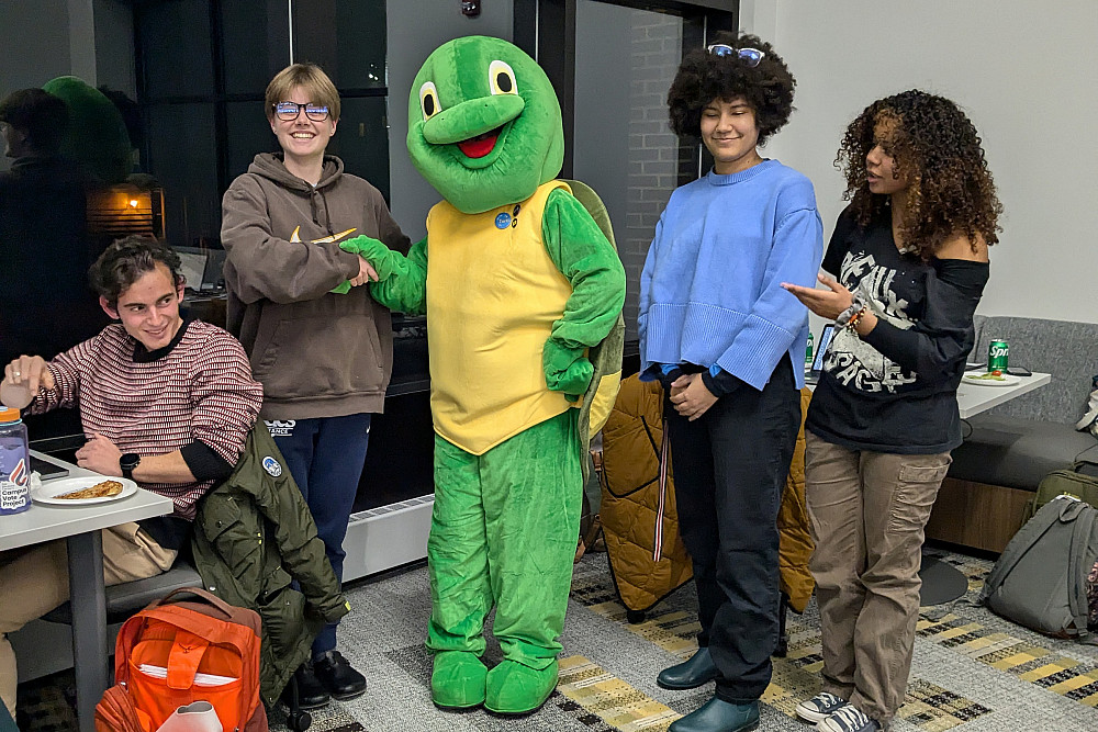 PXL 20241206 031714842.MP Four students smiling and standing with Torti the LITS turtle mascot. One student is shaking Torti's hand.