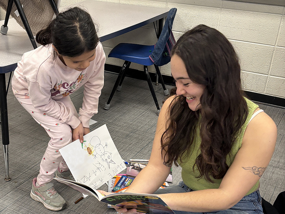 Ella Aizeki reads to a student on the floor of a classroom.