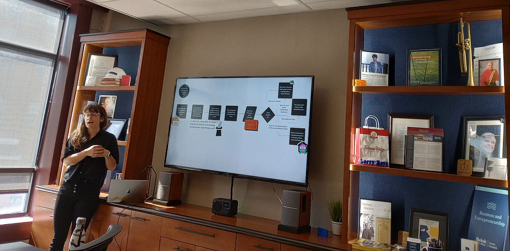 A woman in black stands in front of a TV screen with a presentation and display shelves.