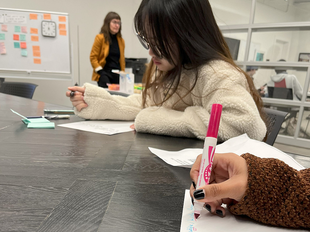 Students are sitting at a table working, while the facilitator stands behind.