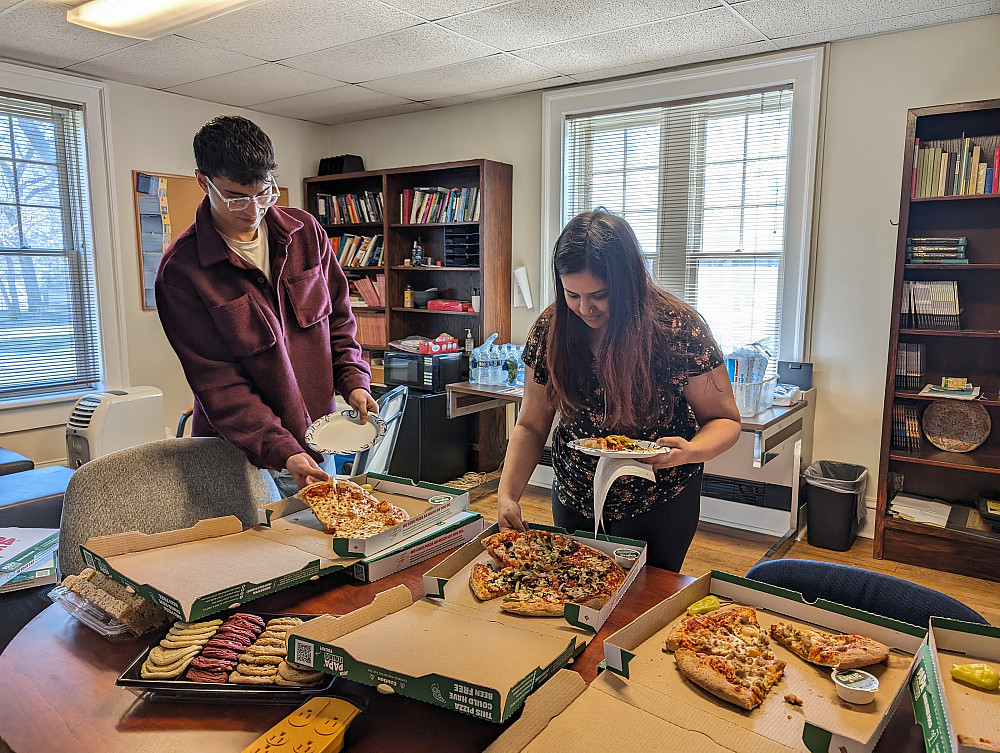 two students stand by a table reaching for pizza