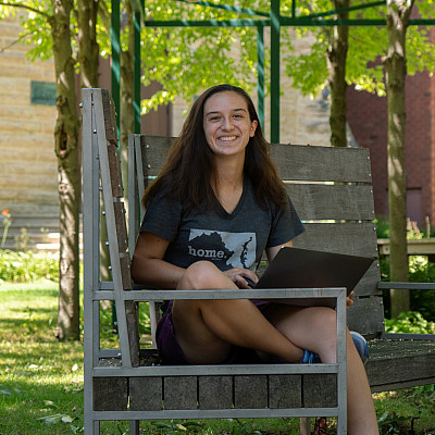 student sits on a bench in the poetry garden with a laptop and green foliage around