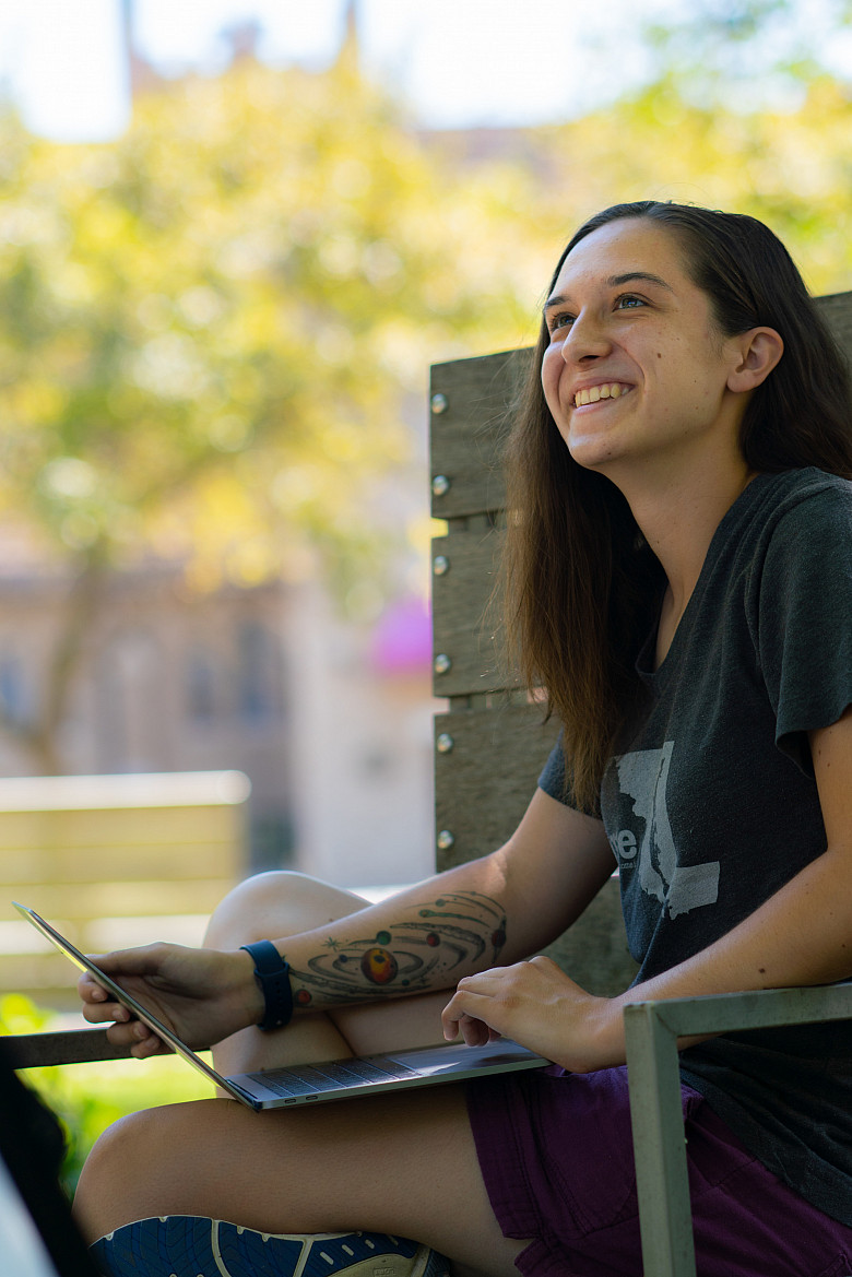 student sits on a bench in the poetry garden with a laptop and green foliage around