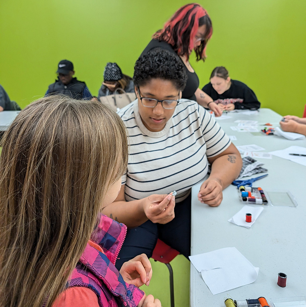 a person holds a needle threader device while a young attendee looks on.
