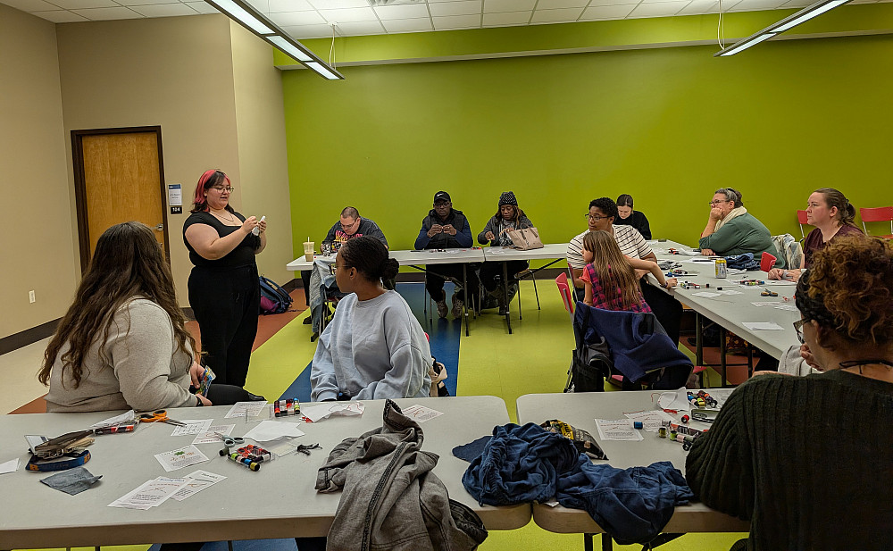 A person stands in front of tables of attendees showing fabric stitches.