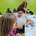 a person holds a needle threader device while a young attendee looks on.