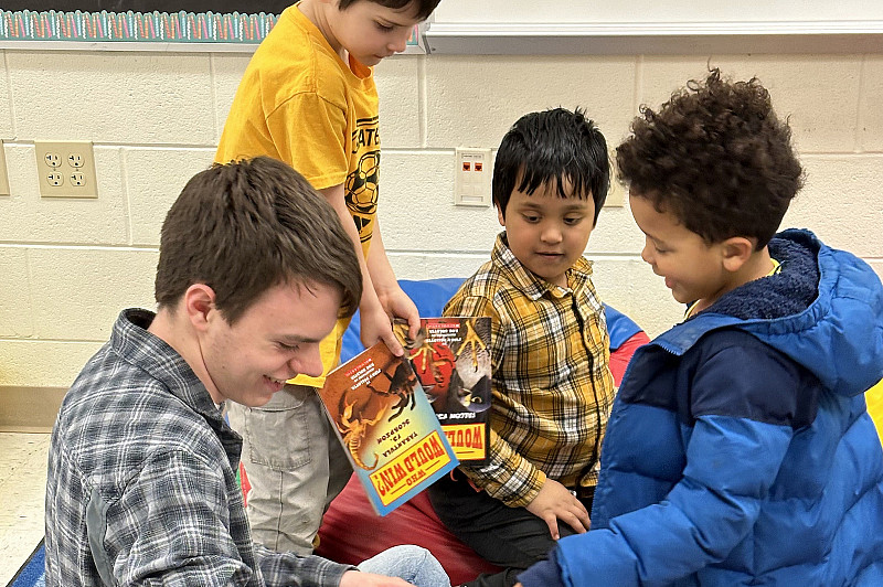 A student is sitting and reading to 3 elementary school students.
