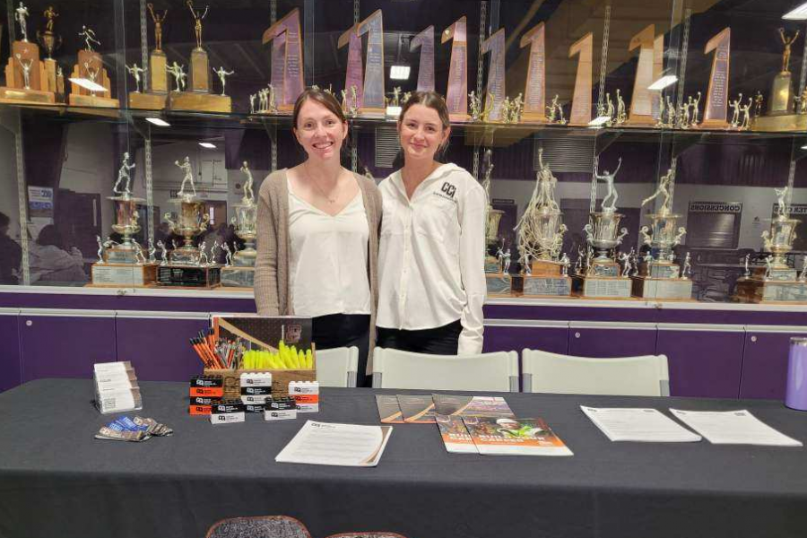 A student and her supervisor stand behind an information table. There are flyers and markers on the table and an awards display behind them.