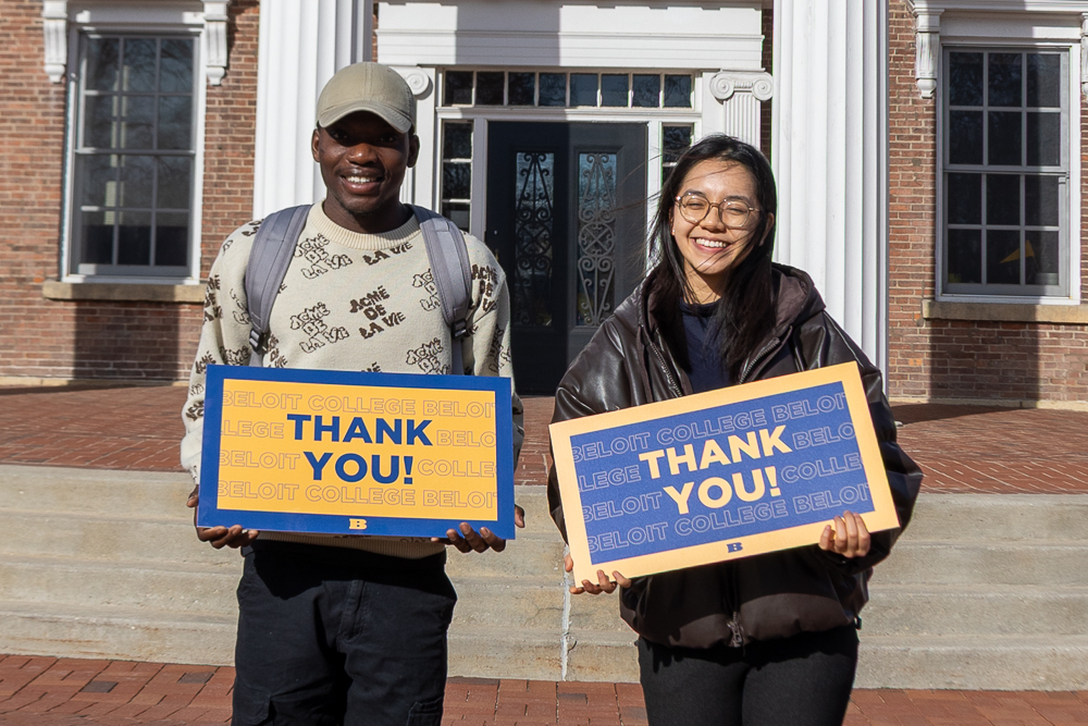Two Beloit College students stand in front of Middle College, smiling, holding signs reading Thank you!