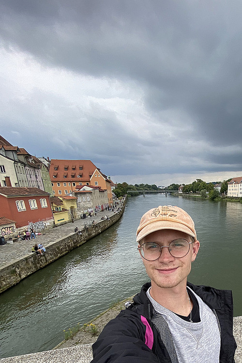 A student in a baseball hat stands on the Old Bridge of Regensburg with the Danube in the background, and foreboding skies in the distance