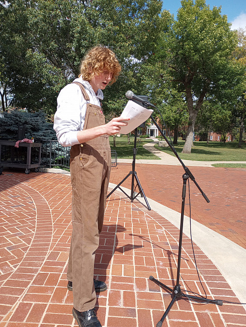 student in overalls and a sweatshirt stands in front of a microphone reading from a photocopy