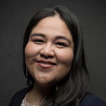 portrait of a Native American woman with long black hair wearing a pink shirt and black blazer while smiling on a dark gray background
