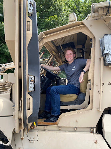 AJ sits in the driver's seat of a semi-truck, smiling at a camera out the open side door