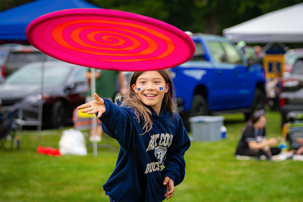 A young child in decked in Beloit College swag throws a giant pink Frisbee towards the viewer, with a giant smile.