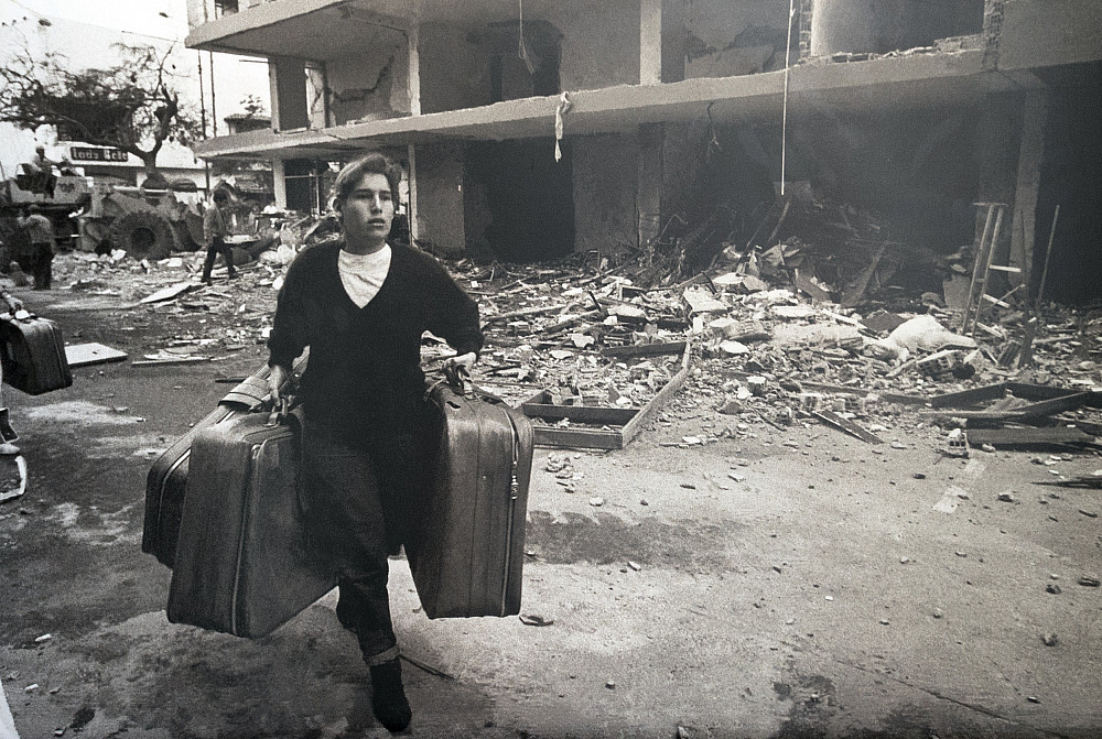 A person runs with three bags of luggage through ruined buildings.